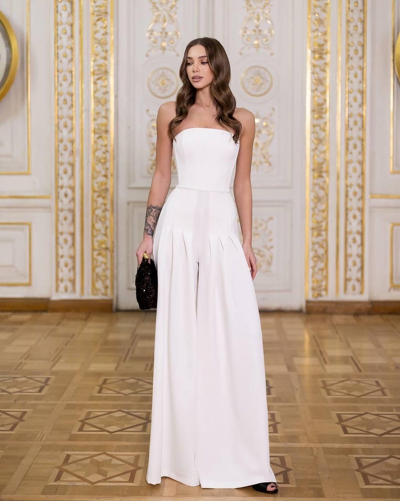 May include: A strapless white jumpsuit with wide-leg pants. The outfit features a fitted bodice and a thin belt at the waist. The person is holding a black sequined clutch. The background is a room with ornate gold trim and a patterned wooden floor.