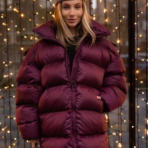Puede incluir: Una chaqueta acolchada burdeos con cuello alto y cierre de cremallera. La chaqueta se lleva con una boina gris. El fondo presenta una cadena de luces blancas cálidas.