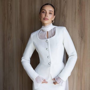 May include: A white suit featuring a tailored jacket with decorative buttons and a high collar with a sheer, polka-dot insert. The suit includes a matching skirt and is set against a wooden background. The jacket has long sleeves with sheer, polka-dot cuffs.