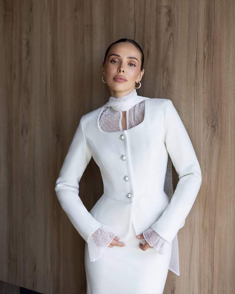 May include: A white suit featuring a tailored jacket with decorative buttons and a high collar with a sheer, polka-dot insert. The suit includes a matching skirt and is set against a wooden background. The jacket has long sleeves with sheer, polka-dot cuffs.
