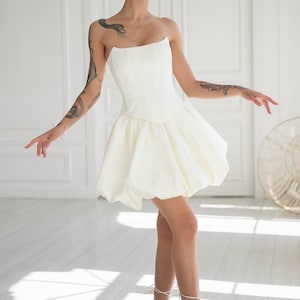 May include: A strapless white mini dress with a fitted bodice and a bubble skirt. The dress is paired with white strappy heels. The model has short blonde hair and tattoos on their arms. The background is a bright, minimalist interior with white walls and a door.