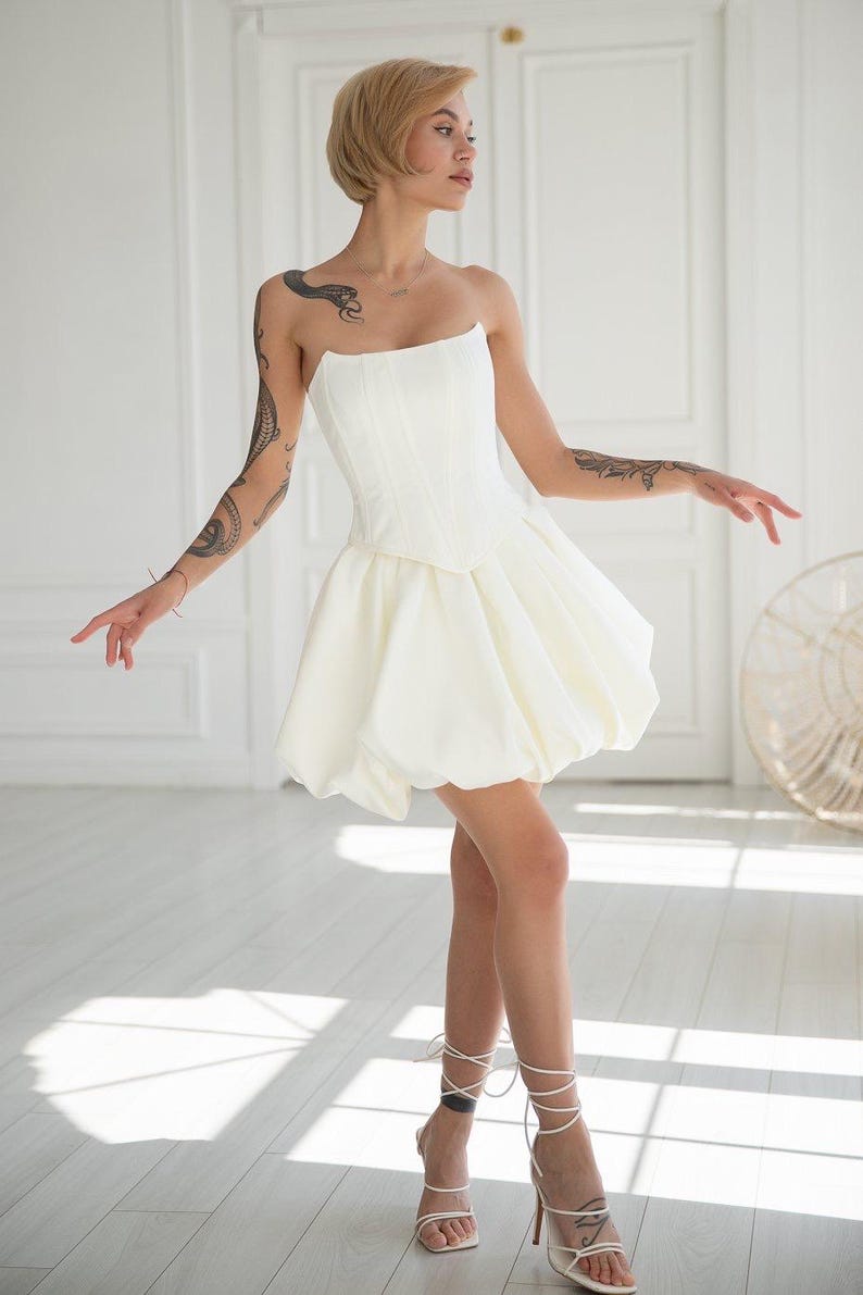 May include: A strapless white mini dress with a fitted bodice and a bubble skirt. The dress is paired with white strappy heels. The model has short blonde hair and tattoos on their arms. The background is a bright, minimalist interior with white walls and a door.