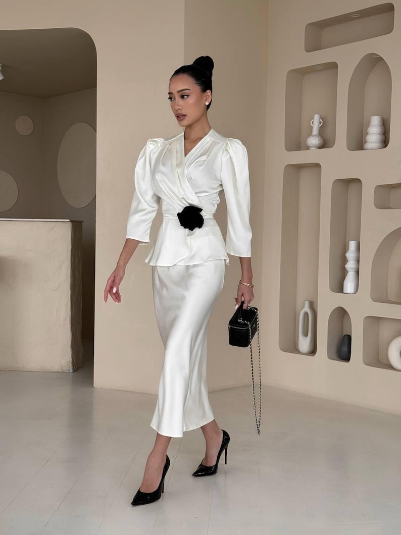 May include: A woman wearing a white satin wrap top with a black flower accent at the waist and a matching white satin skirt. She is wearing black heels and carrying a small black purse.