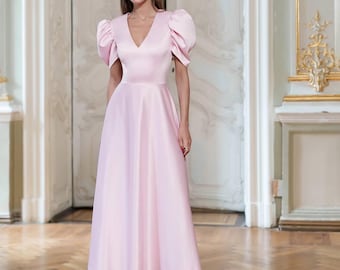 Könnte beinhalten: Ein hellrosa Satin-Kleid mit V-Ausschnitt und puffärmeligen Ärmeln in Ellenbogenlänge. Das Kleid hat ein figurbetontes Oberteil und einen ausgestellten Rock, der bis zur Mitte der Wade reicht. Die Person trägt dunkelbraune High-Heel-Sandalen. Der Hintergrund ist ein Raum mit hellen Wänden und Parkettboden.