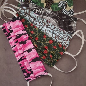 May include: Five fabric face masks with different patterns. The masks are pink and black camouflage, green with red flowers, blue and brown floral, brown and white tie-dye, and green and black animal print.