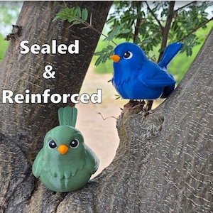 May include: Two decorative bird figurines, one blue and one green, perched on a tree branch. The blue bird has a curved tail and orange beak. The green bird has a small tail and orange beak. The text "Sealed & Reinforced" is visible.