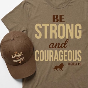 Puede incluir: Una camiseta y gorra de béisbol marrones con la frase "BE STRONG and COURAGEOUS". La camiseta muestra el texto en gran formato, un diseño de león y el versículo bíblico Josué 1:9. La gorra tiene el mismo texto.