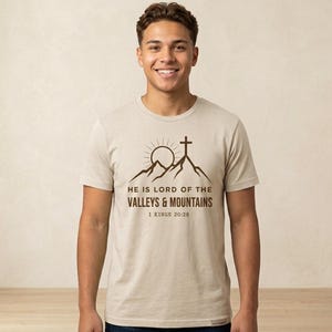 Puede incluir: Una camiseta beige de manga corta con un gráfico marrón oscuro de una cordillera, un sol y una cruz. El texto dice: HE IS LORD OF THE VALLEYS & MOUNTAINS 1 KINGS 20:28. La camiseta tiene cuello redondo y un corte estándar.