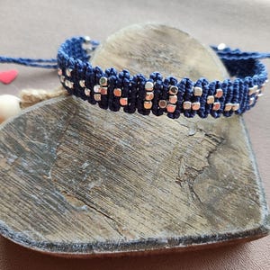 May include: A navy blue friendship bracelet with silver beads. The bracelet is tied in a knot and is lying on a wooden heart.