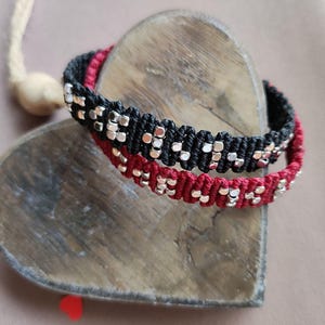 May include: Two friendship bracelets with silver beads woven into black and red cords.