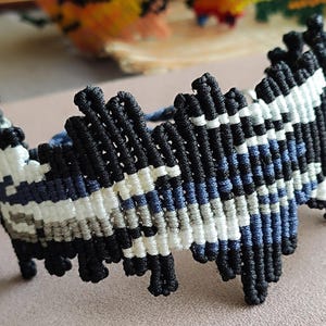 May include: A macrame bracelet featuring a geometric design in black, white, gray, and blue. The bracelet is made of knotted cords, with a textured surface. The bracelet is a unique accessory.
