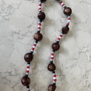 May include: A handmade necklace featuring large, dark brown buckeye nuts interspersed with red, white, black, and gray seed beads. The necklace is displayed against a white and gray marble-patterned surface.