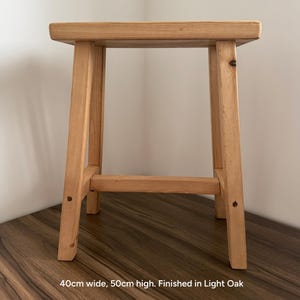 A light oak wooden stool on a dark wood surface against a white wall with text reading 40cm wide, 50cm high. Finished in Light Oak.