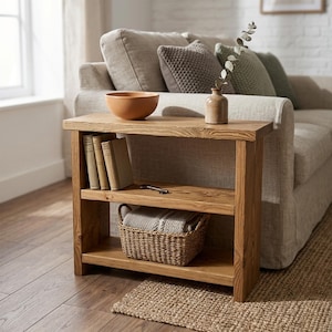 May include: A rectangular wooden console table with two shelves. The top surface holds a terracotta bowl and a small vase with a sprig. The middle shelf contains three books and a metal key. The bottom shelf holds a woven basket with a textile.