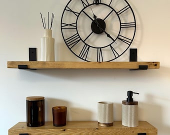 May include: Two natural wood floating shelves with black metal brackets. The top shelf holds a white ribbed reed diffuser and a large black metal wall clock with Roman numerals. The bottom shelf holds a brown glass jar, a brown candle, a ribbed cup, and a soap dispenser.