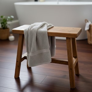 May include: A small wooden stool with a rectangular top and angled legs. A folded grey towel is draped over the stool. The stool is made of light brown wood and is in a bathroom setting.
