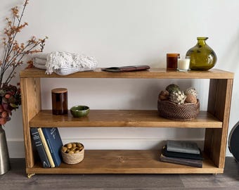May include: A rectangular wooden console table with three horizontal shelves, featuring a medium brown wood grain finish. The unit is supported by small gold-toned casters and holds various decorative items including books, baskets, and glass vessels.