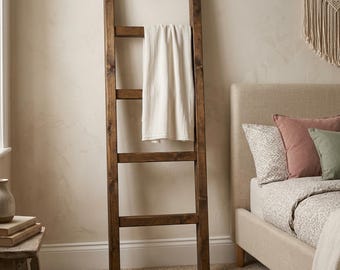 May include: A tall, dark brown wooden ladder with four horizontal rungs, leaning against a wall. A single, plain off-white fabric blanket is draped neatly over the second rung from the top, hanging down to cover the third rung of the wooden structure.