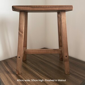 A walnut-finished wooden stool on a dark surface against a white wall with text reading 40cm wide, 50cm high. Finished in Walnut.