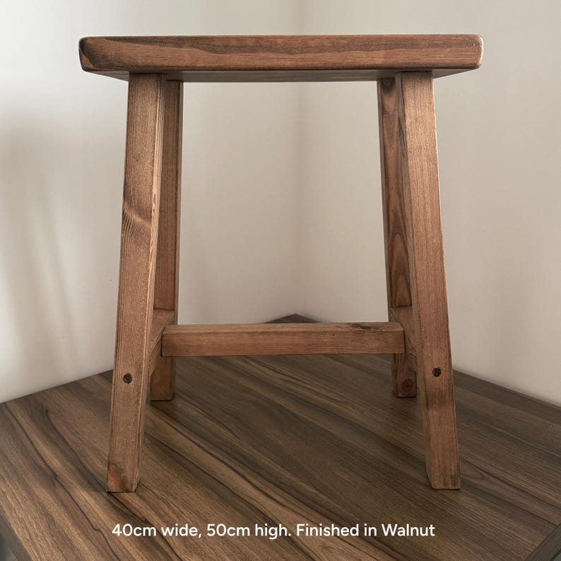 A walnut-finished wooden stool on a dark surface against a white wall with text reading 40cm wide, 50cm high. Finished in Walnut.