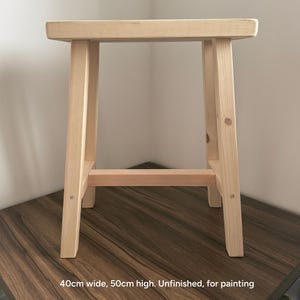 An unfinished light wood stool on a dark surface against a white wall with text reading 40cm wide, 50cm high. Unfinished, for painting.
