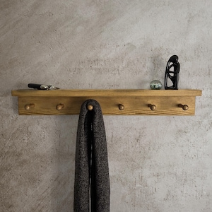 May include: A rectangular wooden wall shelf featuring six rounded pegs along the bottom edge. A dark grey textured scarf hangs from the centre peg. A small black object and a black abstract sculpture sit on the top surface of the wooden shelf.