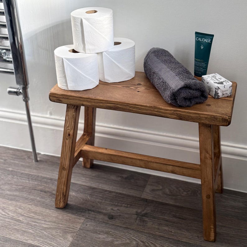 A wooden stool holds three rolls of toilet paper, a folded grey towel, a tube of hand cream, and a bar of soap against a white wall.