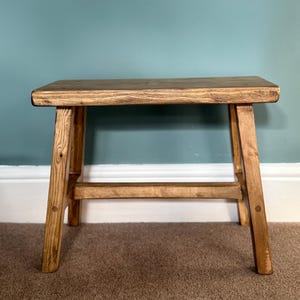 A small wooden stool with splayed legs and a lower support rail stands on a brown carpet against a teal wall.