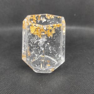 Peut inclure: Un récipient hexagonal transparent en résine incrusté de flocons métalliques dorés et argentés. L'objet présente une finition lisse et translucide avec une forme géométrique marquée et une large ouverture supérieure pour ranger de petits objets.