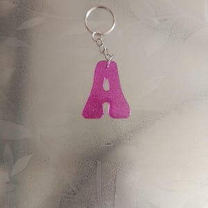 Peut inclure: Un porte-clés en forme de lettre A rose vif avec une finition pailletée. La lettre est fixée à un anneau brisé en métal argenté par une courte chaîne. Le design est lisse et translucide, affichant une teinte magenta vibrante avec des éclats.