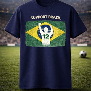 Puede incluir: Camiseta azul marino con el texto "SUPPORT BRAZIL" sobre una bandera brasile&ntilde;a desgastada. El dise&ntilde;o de la bandera incluye una silueta blanca de una persona con los brazos levantados y el n&uacute;mero 12.