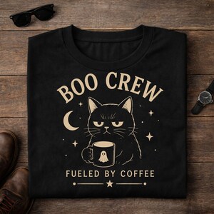 Op de afbeelding: Een zwart t-shirt met een afbeelding van een norse kat die een mok met een spookillustratie vasthoudt. De tekst luidt BOO CREW en FUELED BY COFFEE, vergezeld door een halve maan en sterren in een lichtbeige kleur op de donkere stof.