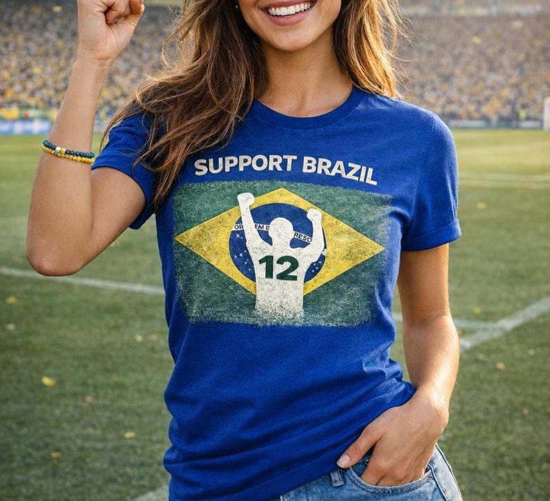 Puede incluir: Una camiseta azul con la frase "SUPPORT BRAZIL" sobre un dise&ntilde;o desgastado de la bandera brasile&ntilde;a. El gr&aacute;fico incluye una silueta de persona con los brazos levantados y el n&uacute;mero "12". La camiseta la lleva una persona.