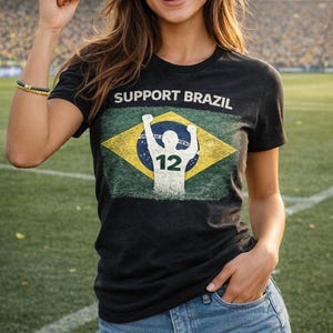 Puede incluir: Camiseta negra con la frase "SUPPORT BRAZIL" sobre un dise&ntilde;o desgastado de la bandera brasile&ntilde;a. El gr&aacute;fico de la bandera presenta una silueta blanca de una persona con los brazos levantados y el n&uacute;mero 12.
