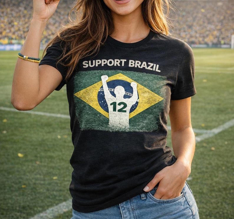 Puede incluir: Camiseta negra con la frase "SUPPORT BRAZIL" sobre un dise&ntilde;o desgastado de la bandera brasile&ntilde;a. El gr&aacute;fico de la bandera presenta una silueta blanca de una persona con los brazos levantados y el n&uacute;mero 12.