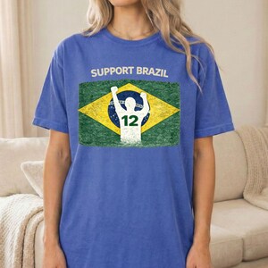 Puede incluir: Camiseta azul con el texto "SUPPORT BRAZIL" sobre un gr&aacute;fico de la bandera brasile&ntilde;a. El dise&ntilde;o de la bandera incluye la silueta de una persona con los brazos levantados y el n&uacute;mero 12. La camiseta es de color azul liso.