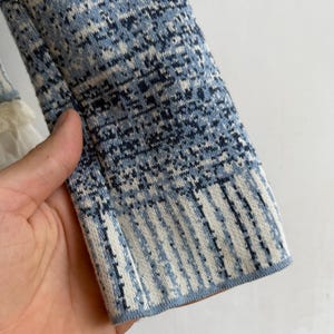 May include: A long-sleeved knit garment featuring a textured pattern in shades of blue, navy, and white. The sleeve displays a mottled, speckled design on the upper portion, transitioning into a vertical striped pattern at the ribbed cuff edge.