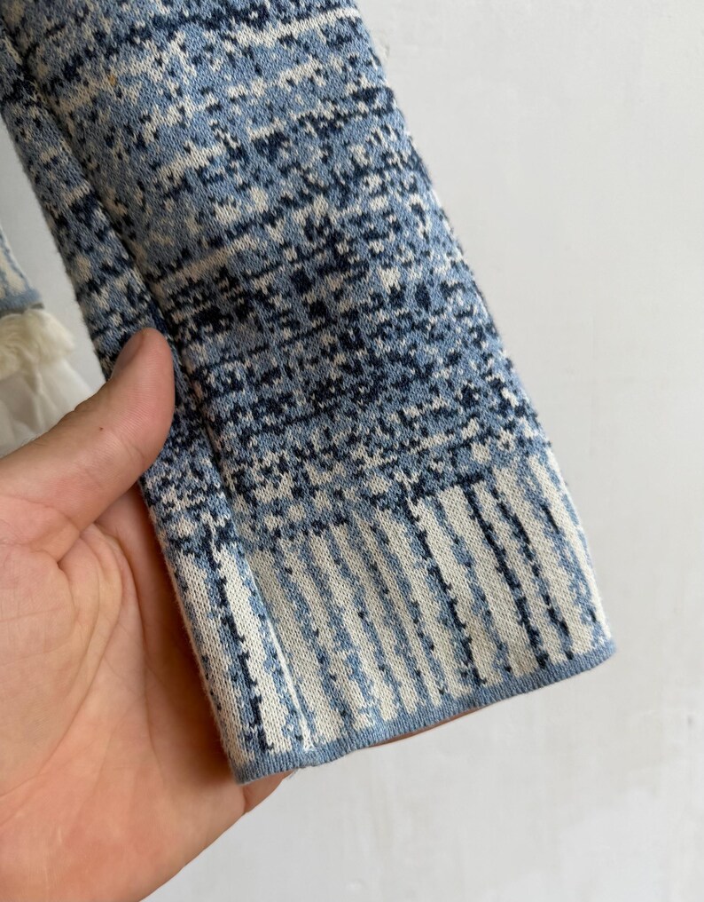 May include: A long-sleeved knit garment featuring a textured pattern in shades of blue, navy, and white. The sleeve displays a mottled, speckled design on the upper portion, transitioning into a vertical striped pattern at the ribbed cuff edge.