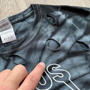 Może przedstawiać: Czarna i szara koszulka tie-dye z dziurami. Koszulka ma biały wz&oacute;r graficzny. Etykieta pokazuje rozmiar M. Koszulka jest wykonana z bawełny i poliestru.