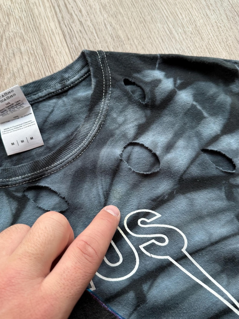 Może przedstawiać: Czarna i szara koszulka tie-dye z dziurami. Koszulka ma biały wz&oacute;r graficzny. Etykieta pokazuje rozmiar M. Koszulka jest wykonana z bawełny i poliestru.