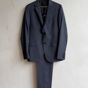 May include: A dark blue suit consisting of a single-breasted blazer with notched lapels, two front buttons, and matching trousers. The blazer features a chest pocket, two flap pockets, and four buttons on each sleeve cuff, all presented on a hanger.