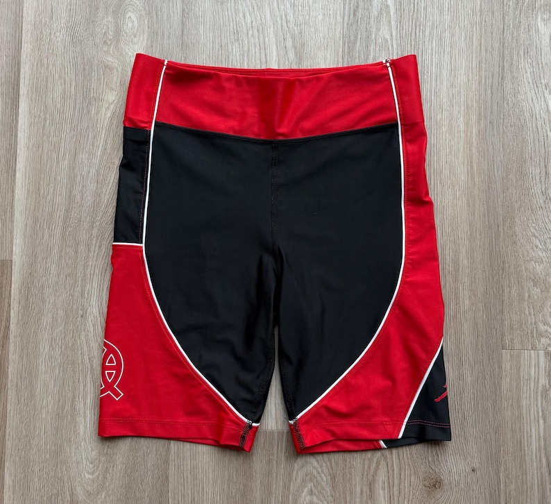 May include: Black and red athletic shorts with white trim. The shorts feature a high waistband and a logo on the left leg. Made from breathable, athletic fabric, these shorts are designed for comfort.