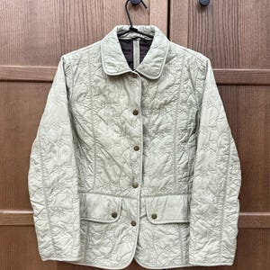May include: A light green quilted jacket with a floral pattern. The jacket has a collared neckline, button closure, and two front pockets. The jacket is hanging on a black hanger.