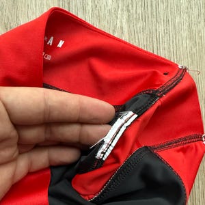 May include: A close-up of a red and black athletic garment. The garment features a red outer layer with black detailing and stitching. The word "RAN" is visible on the red fabric. A hand is holding the garment.