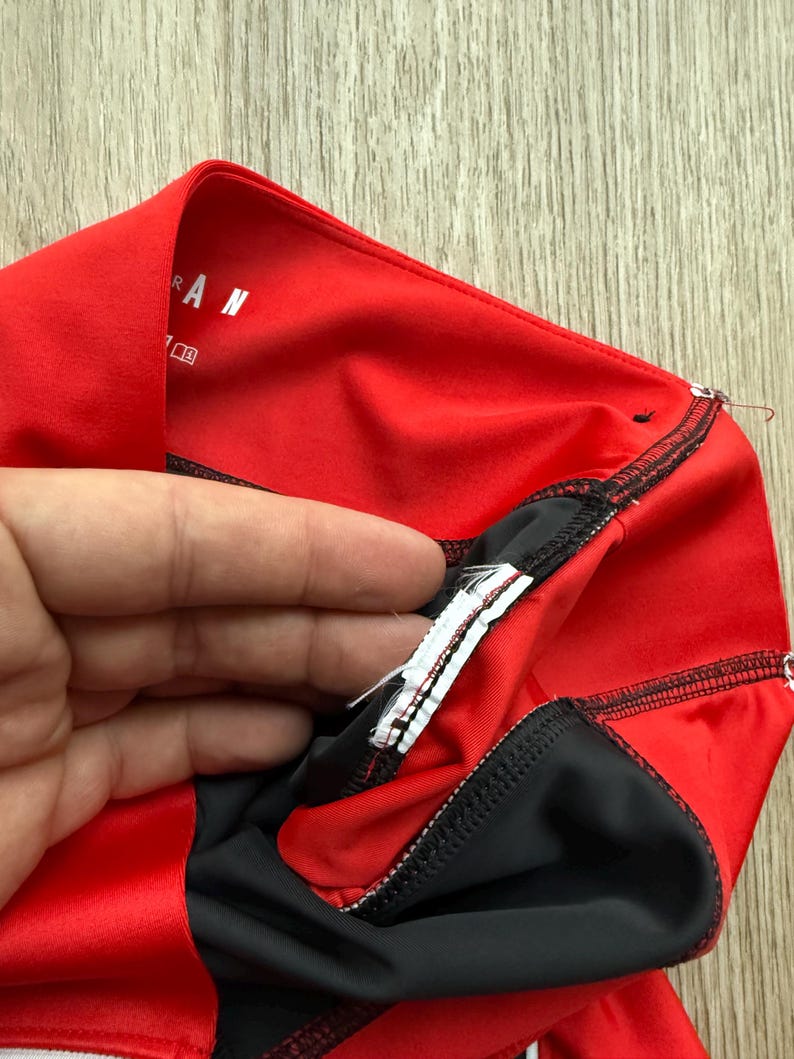 May include: A close-up of a red and black athletic garment. The garment features a red outer layer with black detailing and stitching. The word "RAN" is visible on the red fabric. A hand is holding the garment.