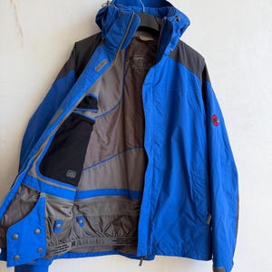 May include: A blue and dark gray hooded jacket with a front zipper closure. The interior lining is gray with black pocket detailing and a powder skirt featuring the Mammut brand name. A red circular logo is visible on the left sleeve.