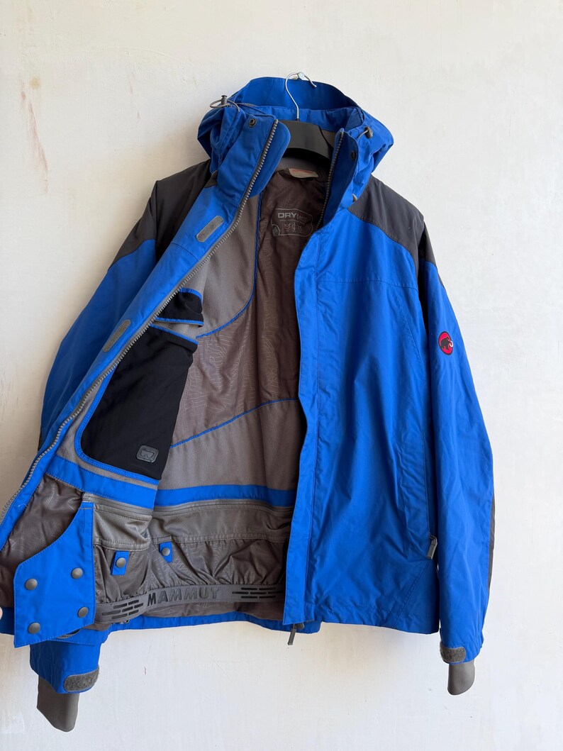 May include: A blue and dark gray hooded jacket with a front zipper closure. The interior lining is gray with black pocket detailing and a powder skirt featuring the Mammut brand name. A red circular logo is visible on the left sleeve.