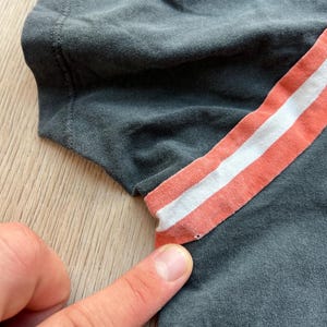 May include: Close-up of a gray t-shirt with a peach and white striped detail on the sleeve.