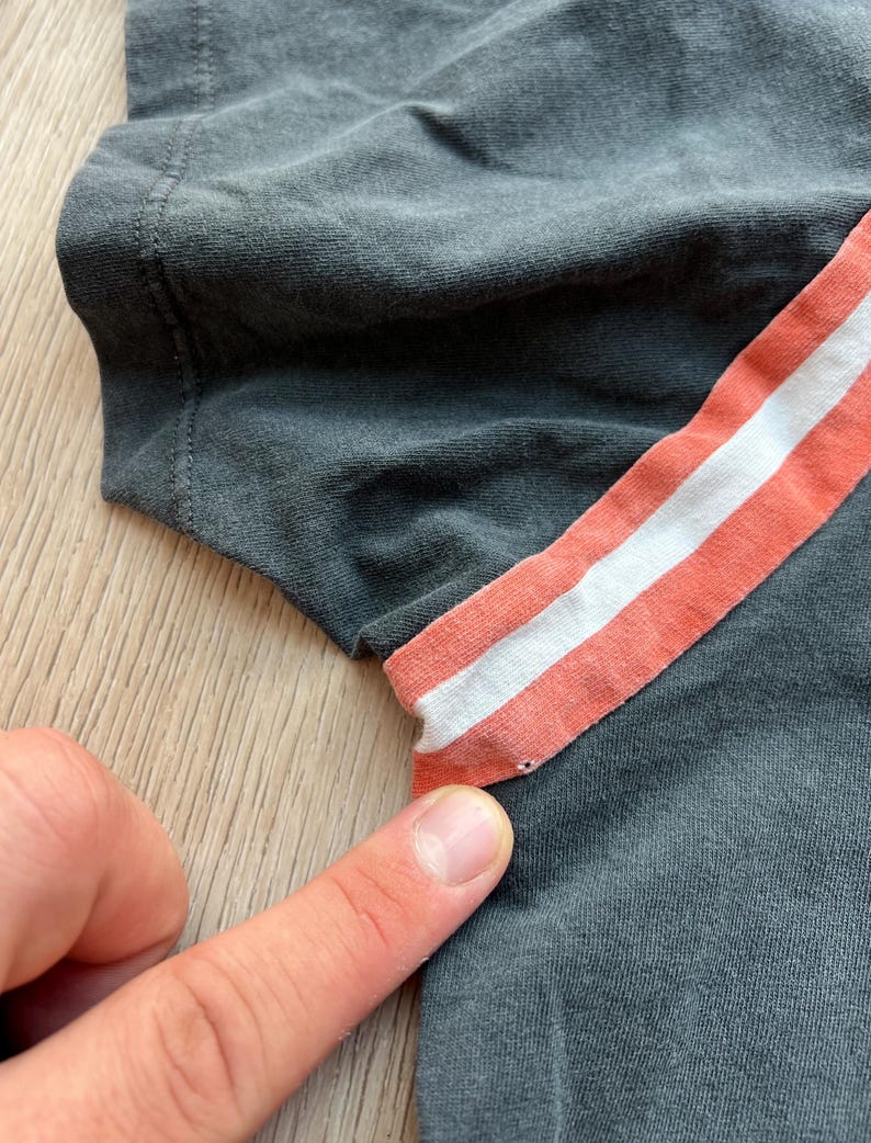 May include: Close-up of a gray t-shirt with a peach and white striped detail on the sleeve.