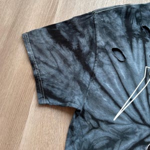 Może przedstawiać: Zbliżenie na koszulkę tie-dye w odcieniach szarości i czerni. Koszulka ma postarzany wygląd z kilkoma małymi dziurami. Białe szwy są widoczne wzdłuż szw&oacute;w rękawa.